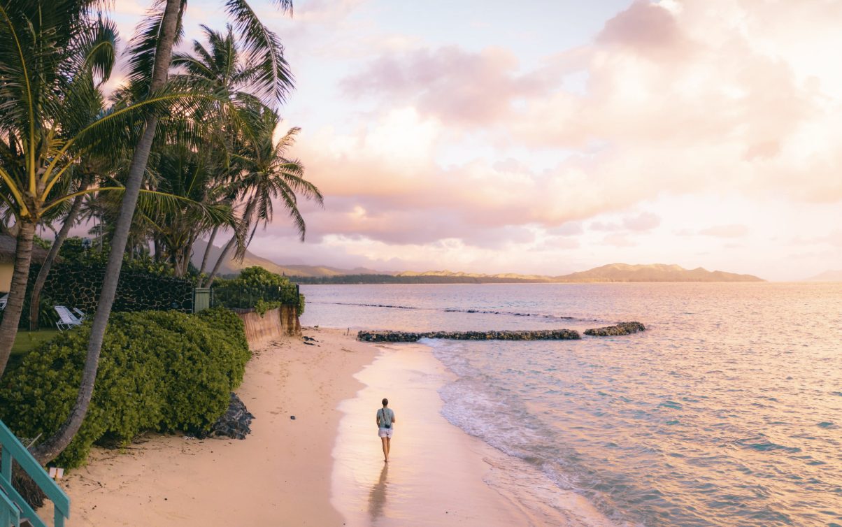 A person walking along a quiet Oahu beach at sunset with palm trees and the Koolau mountains in the distance